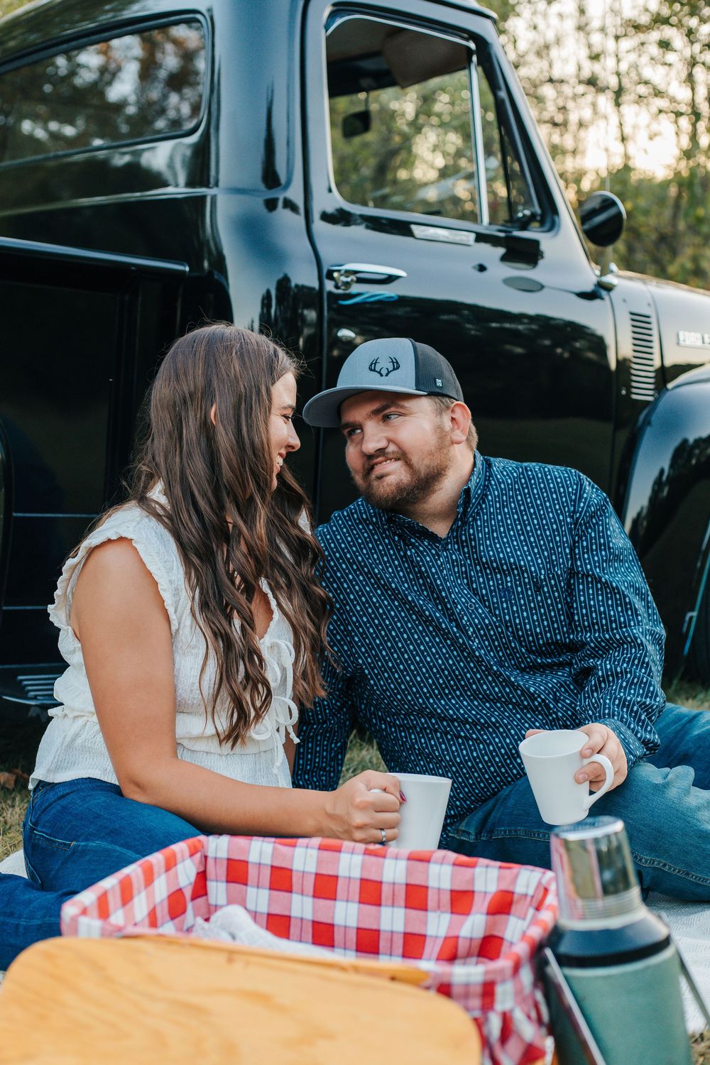 A couple enjoys coffee and a picnic on a red checkered blanket next to a vintage black truck at sunset.