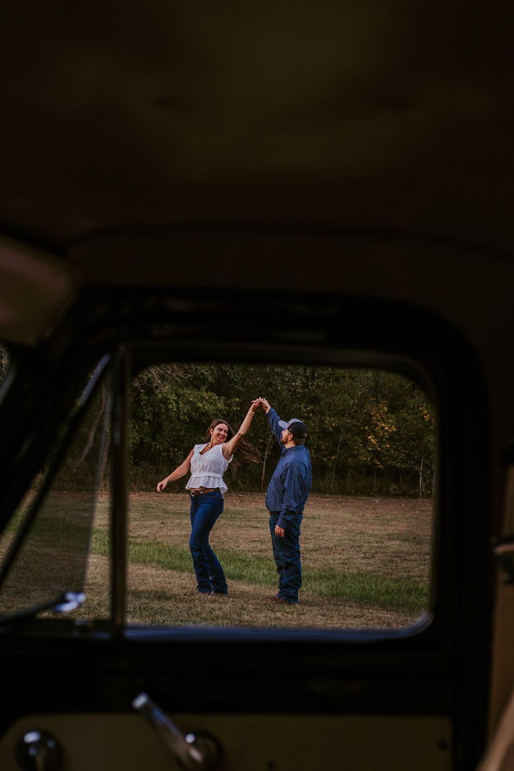 A couple dancing in a field at sunset, viewed through a car window frame.