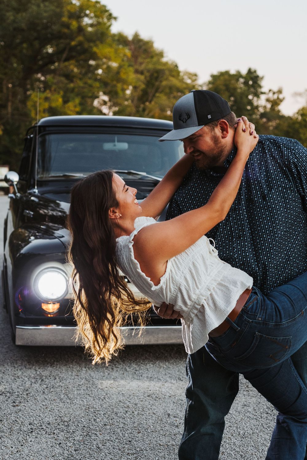A couple dances together in a romantic dip pose at sunset in a parking lot with cars in the background.