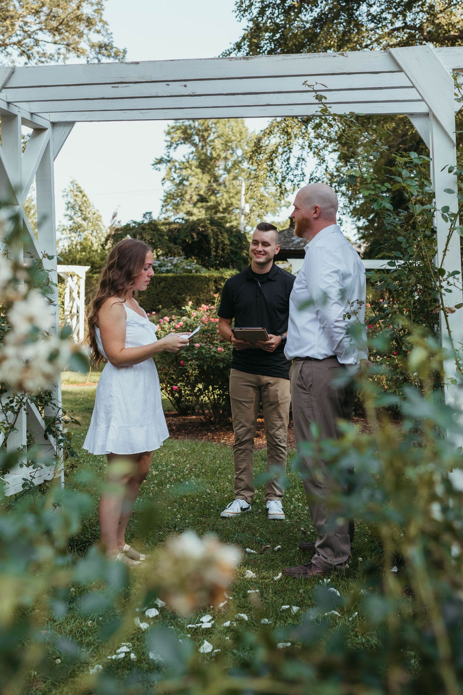 Outdoor wedding ceremony taking place under a white pergola in a garden setting.