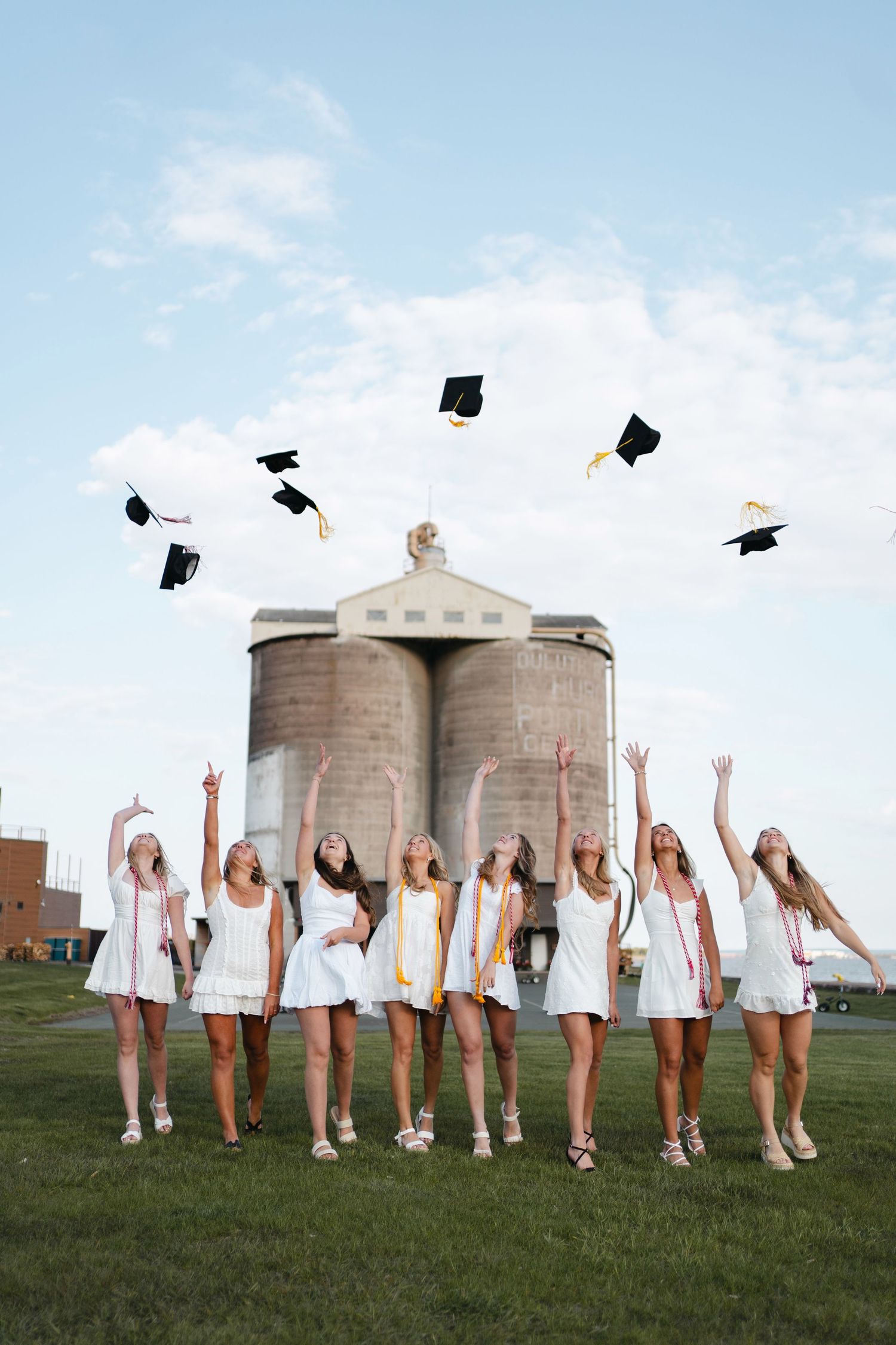 A group in white dresses throws graduation caps in the air while standing in front of industrial storage silos.
