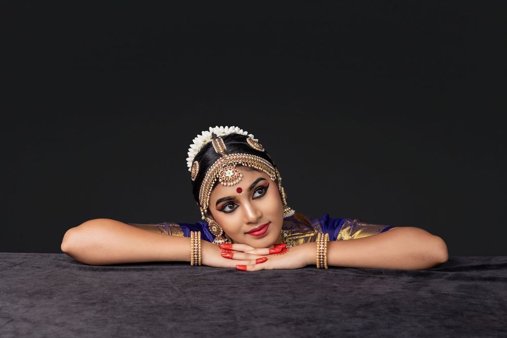 A dancer in traditional Indian dance costume and jewelry rests their head on folded arms against a dark background.