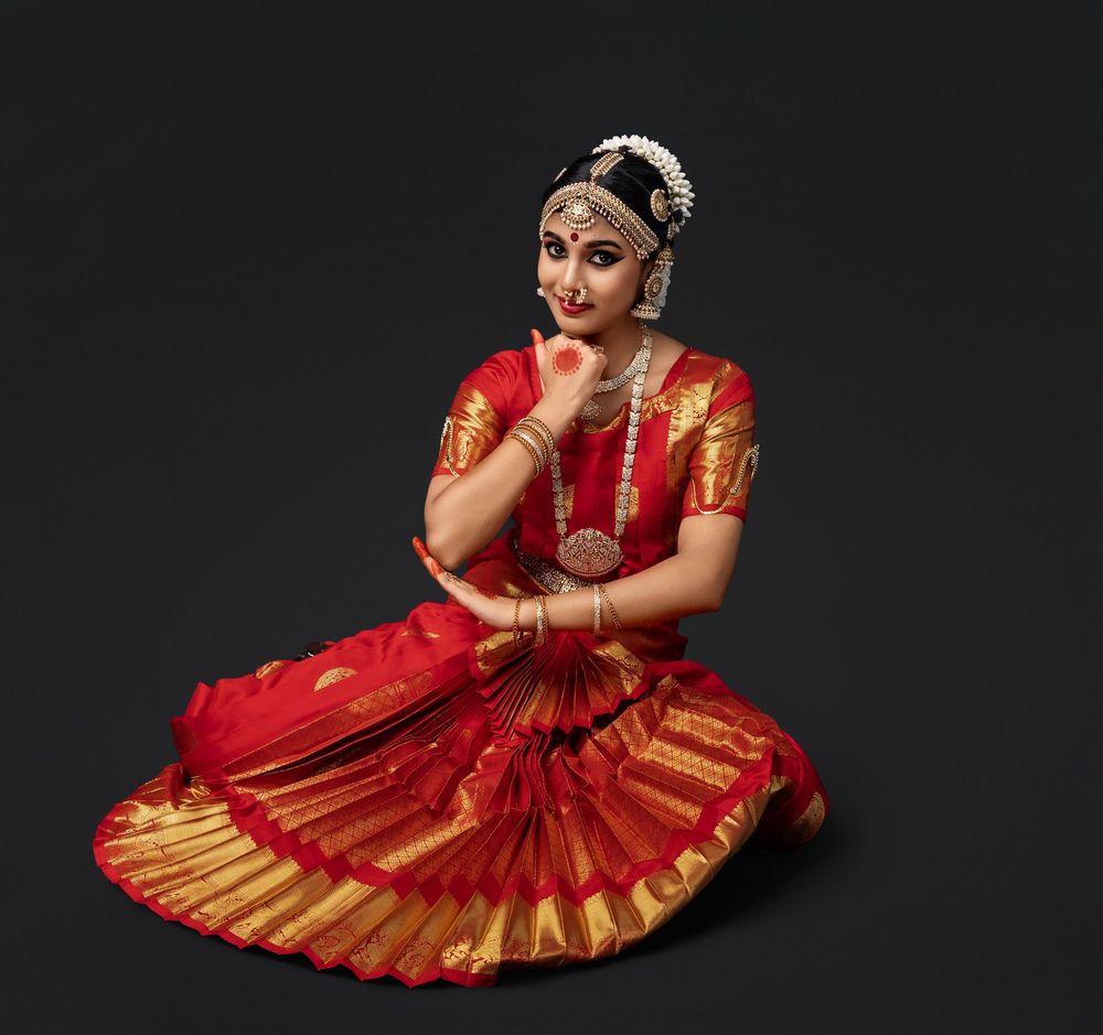 A classical Indian dancer in a red and gold sari performs a traditional pose against a dark background.