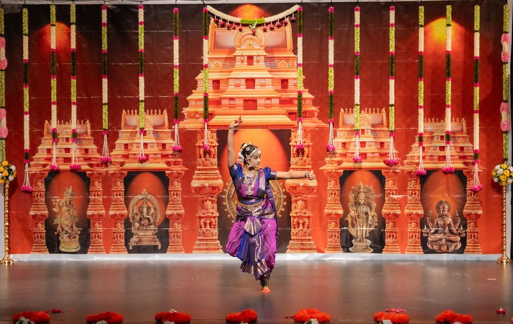 Stage performers dance in traditional costumes in front of an elaborate red and gold temple backdrop with dramatic lighting.