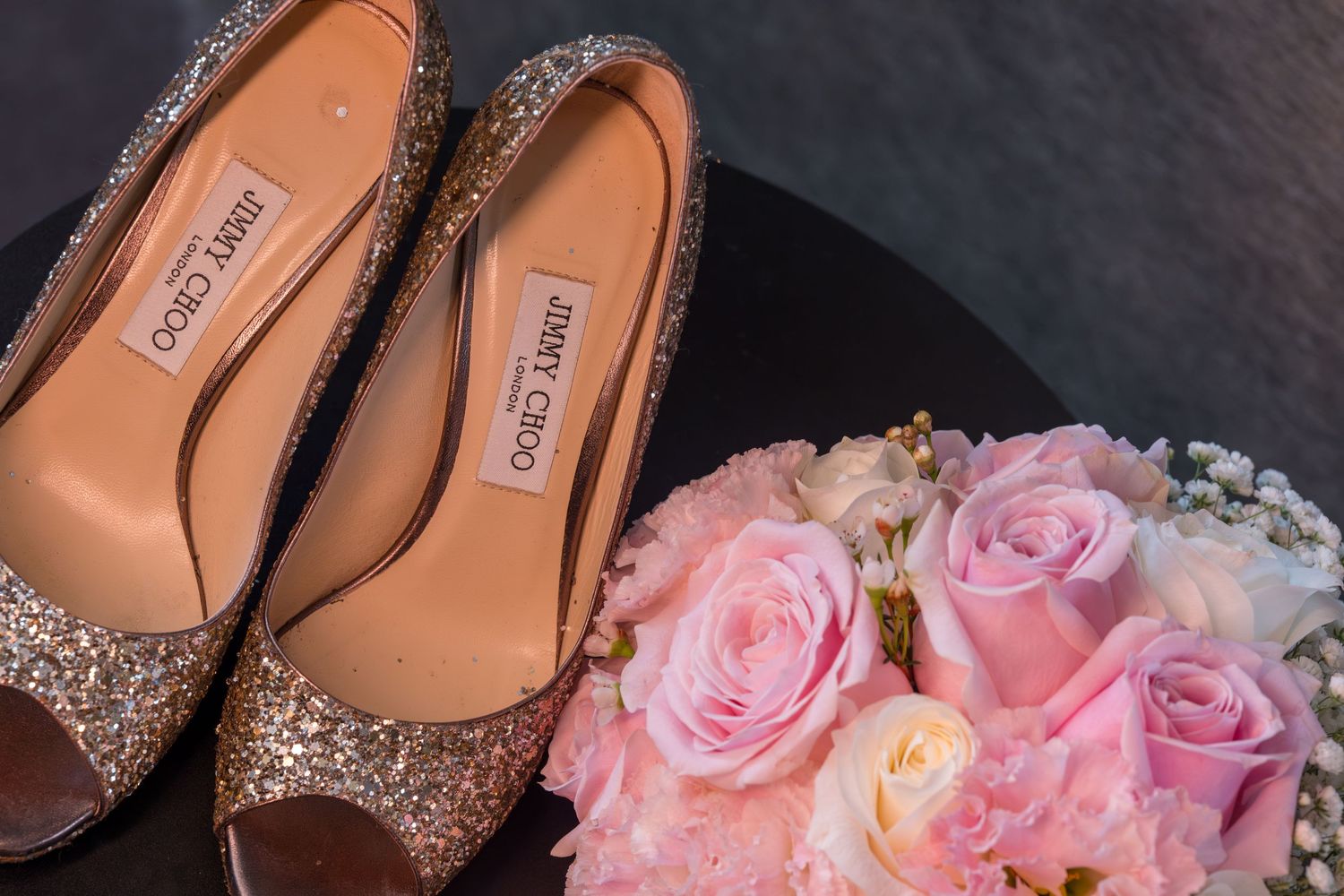 Sparkly gold bridal shoes next to a soft pink rose bouquet against a dark background.