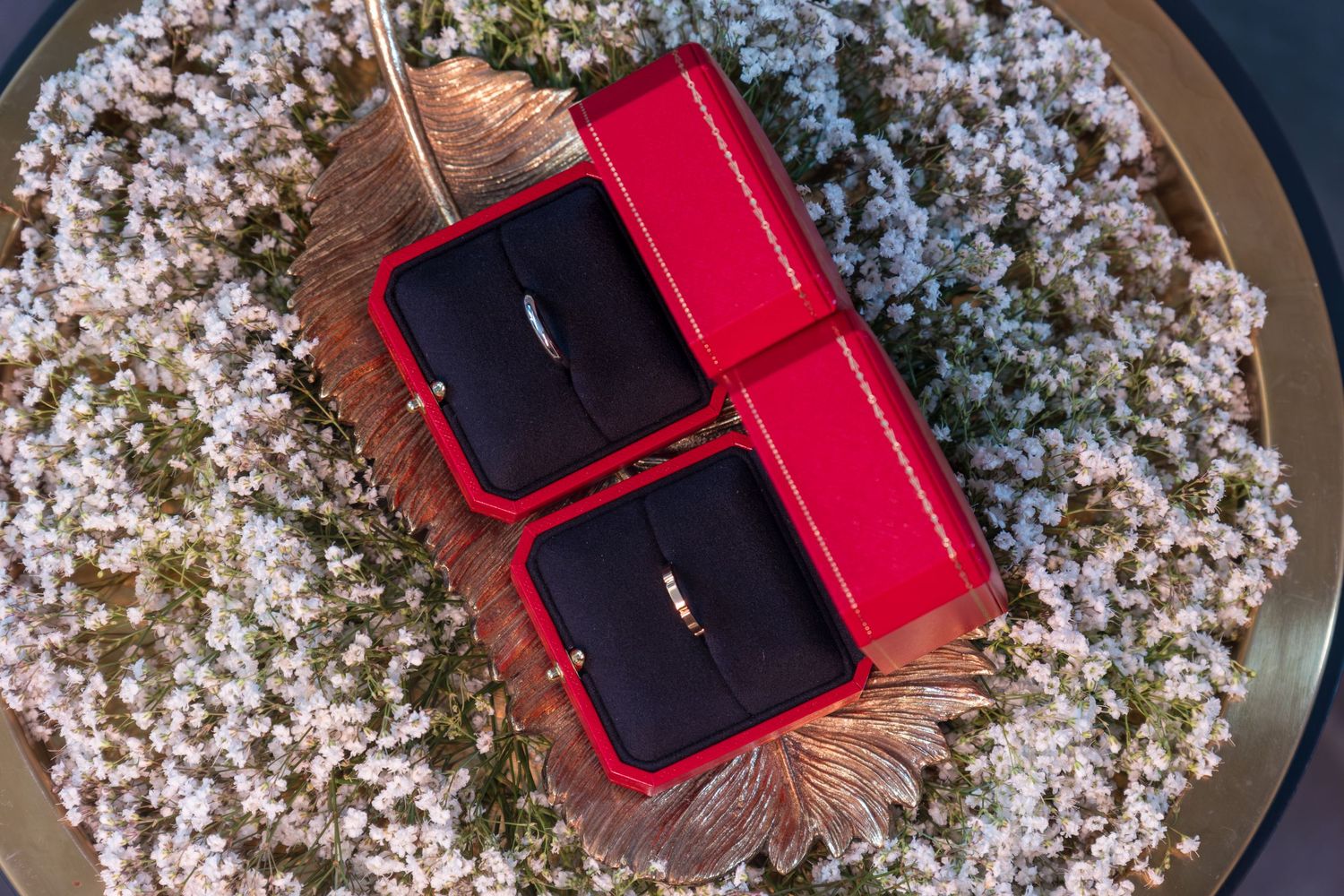Wedding rings in red velvet boxes surrounded by delicate white flower petals on gold tray.