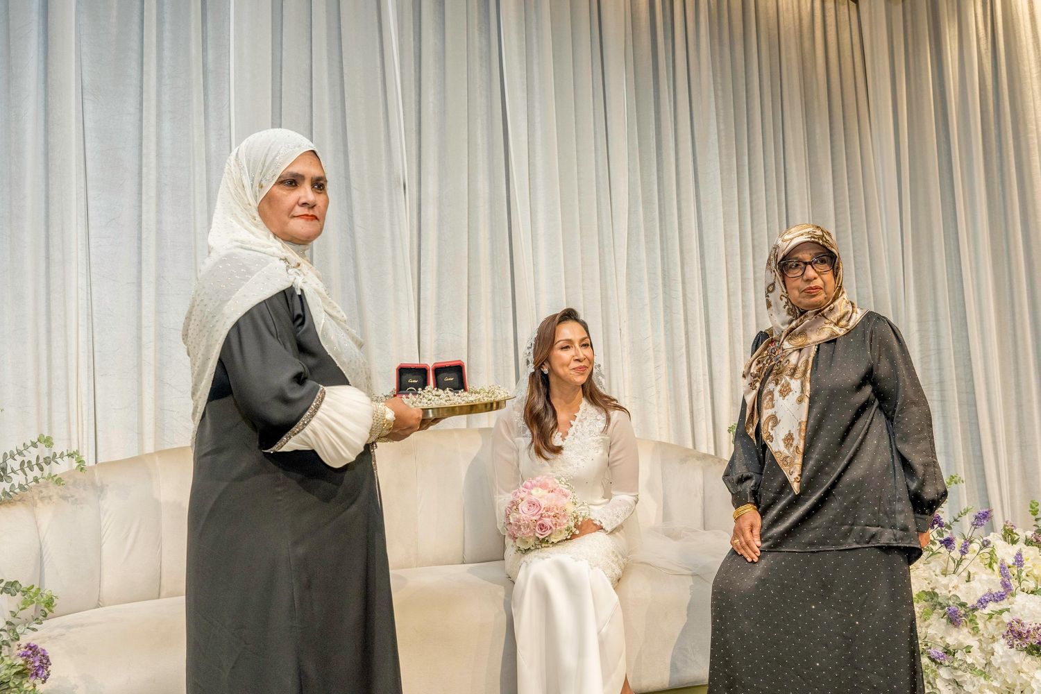 A wedding ceremony celebration with guests in formal attire and hijabs gathered around a decorated table.