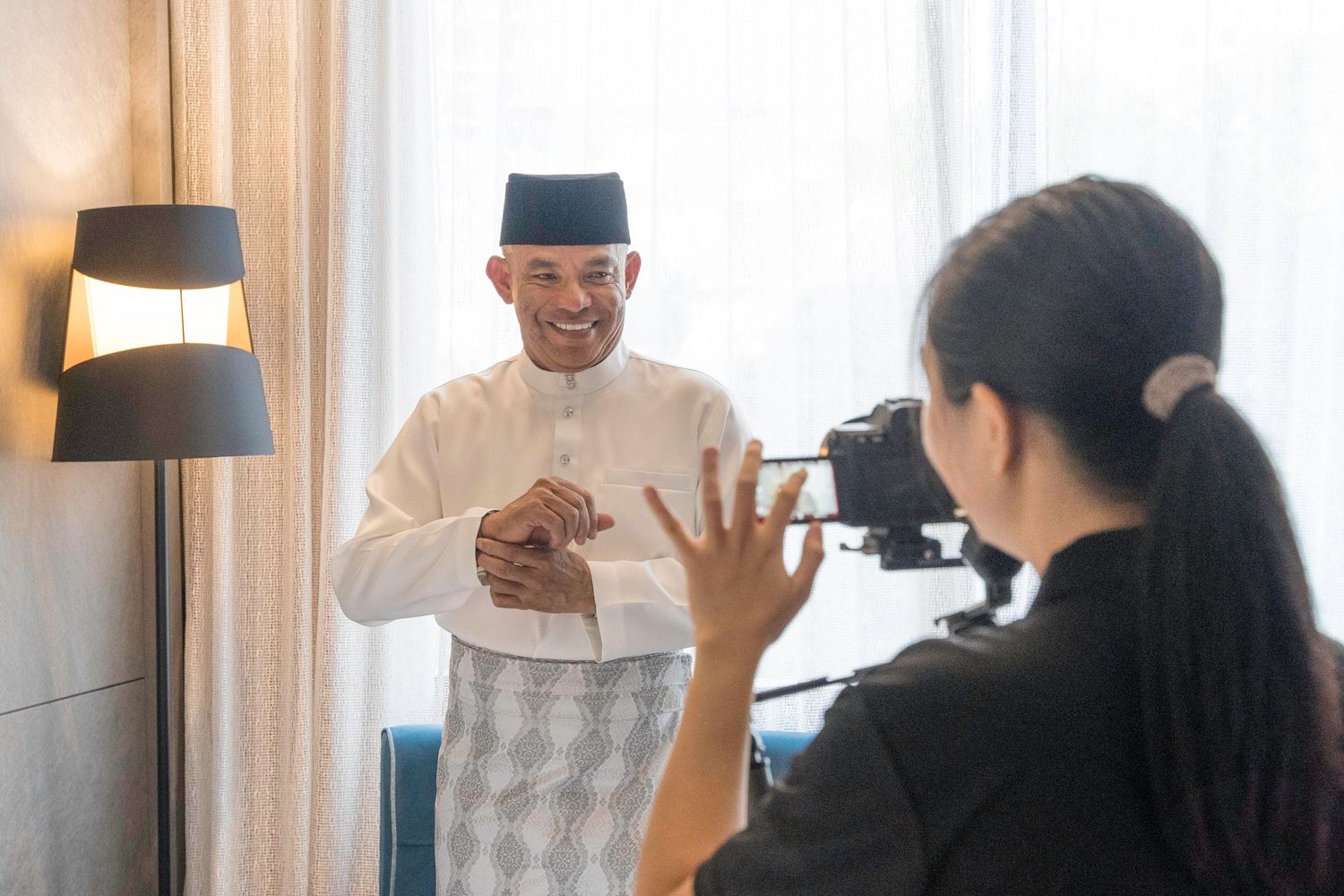 A photographer captures video footage of someone in traditional Malaysian attire speaking in front of a window.