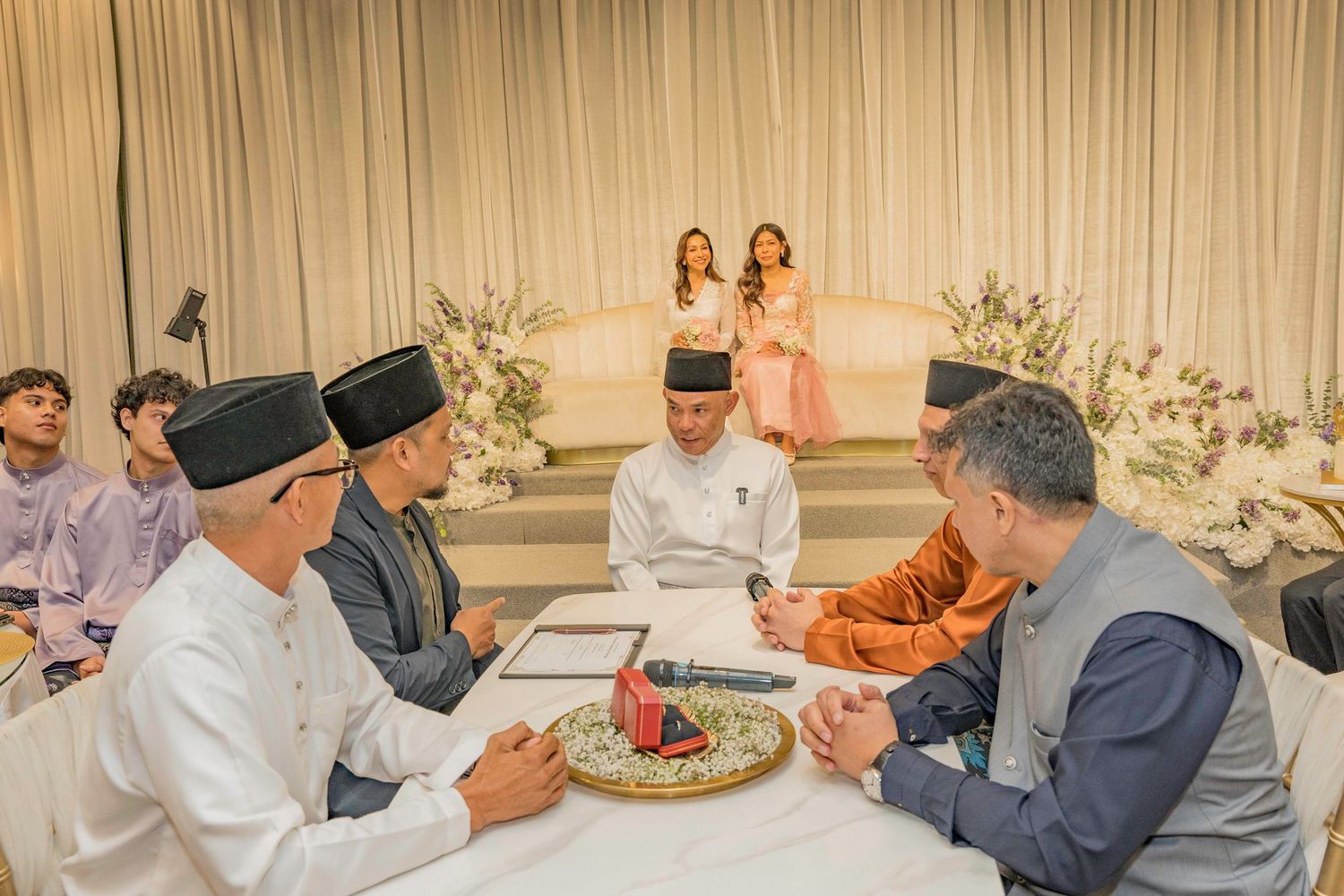 Traditional Malay wedding ceremony with men in songkok hats signing documents at a decorated table.
