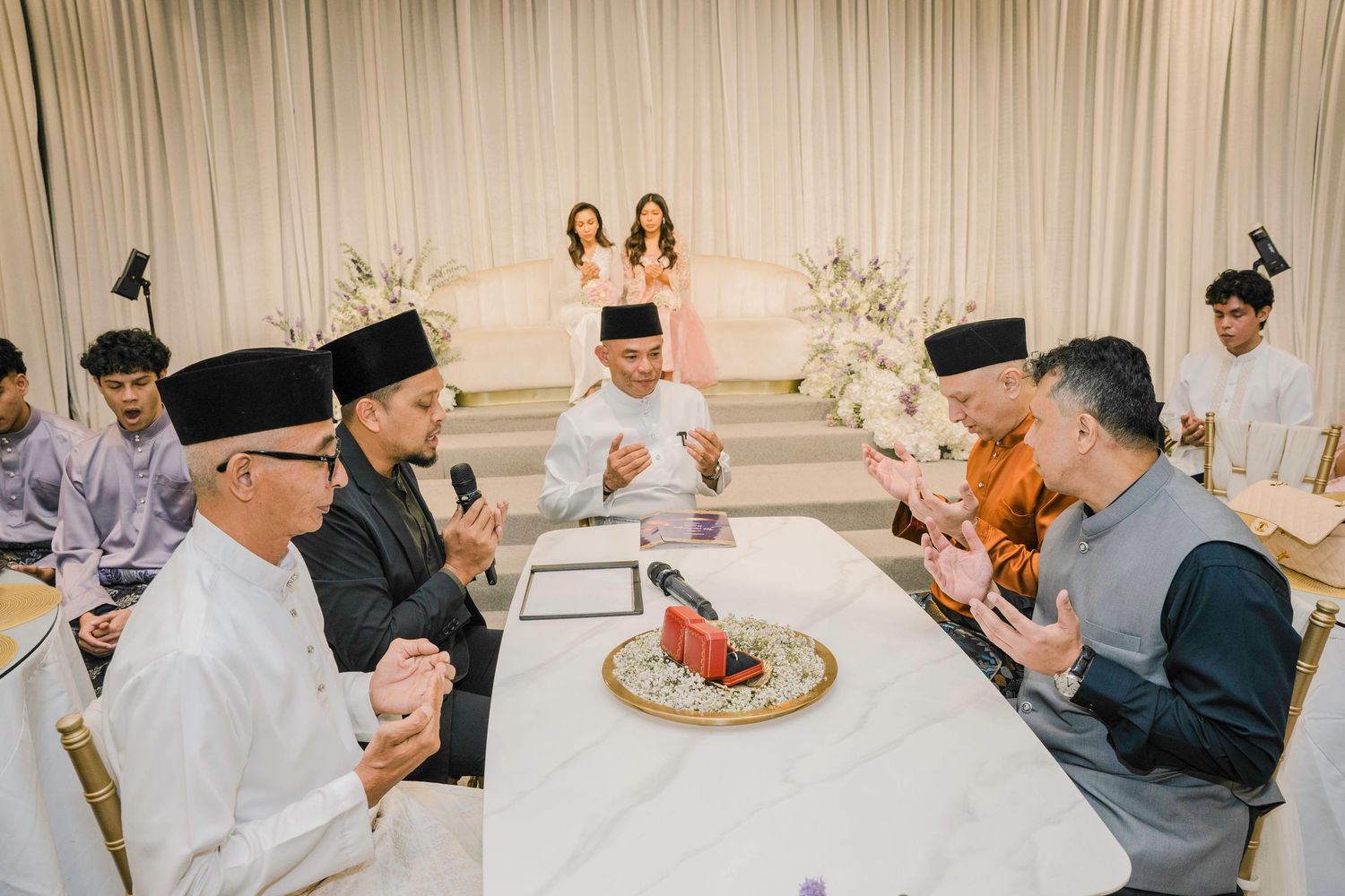 Wedding reception guests gathered around tables in a banquet hall with white floral arrangements.