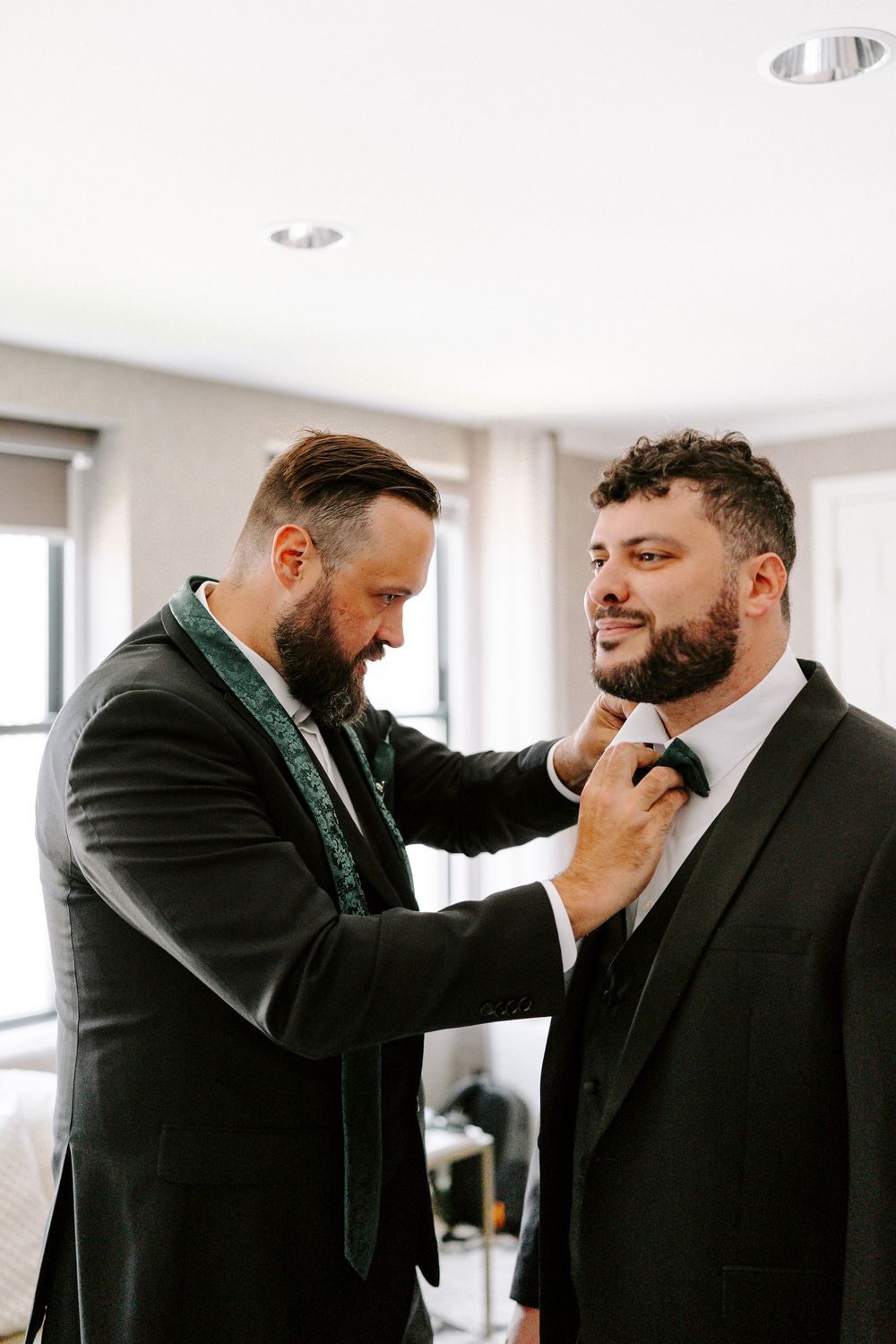 Two people sharing an emotional moment while adjusting formal attire in a bright indoor setting.
