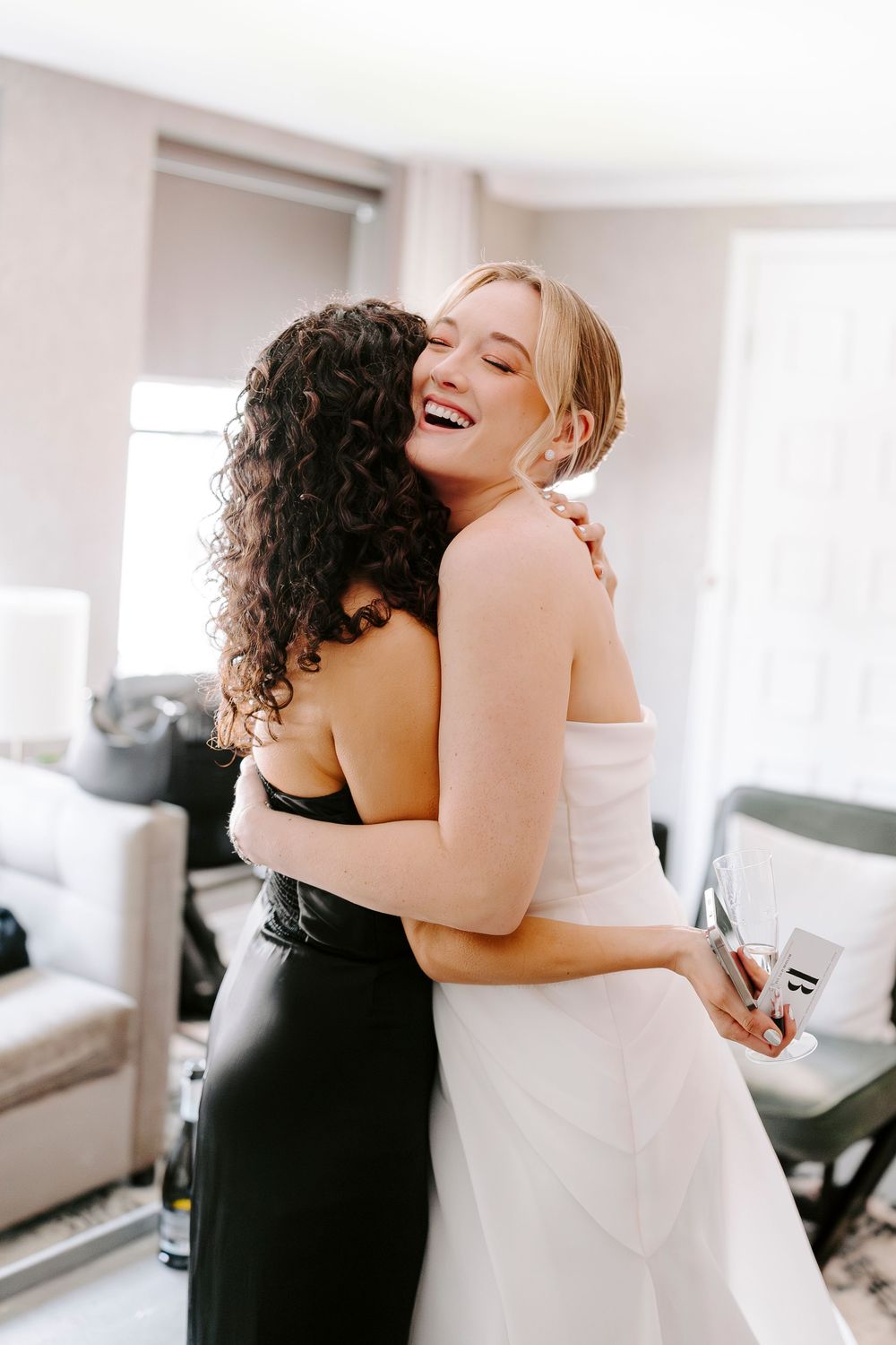 Two people share a joyful embrace in a bright indoor setting, one wearing white and one wearing black.