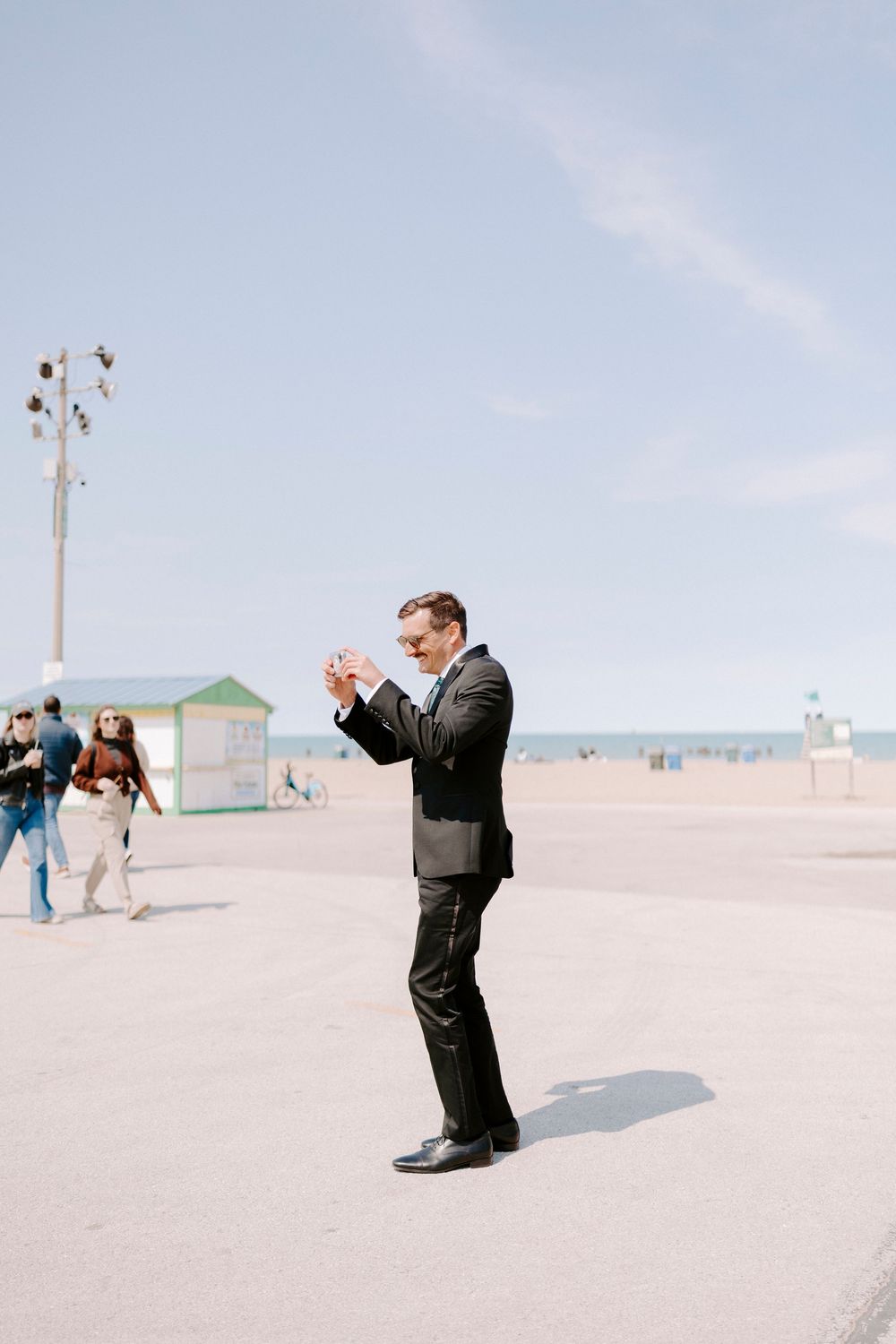 People in formal attire taking photos on a sandy beach with light poles in the background.
