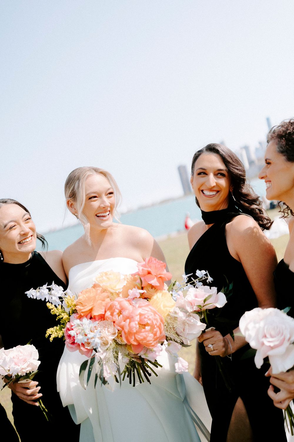 Bridesmaids in black dresses holding coral and white floral bouquets laugh together outdoors.