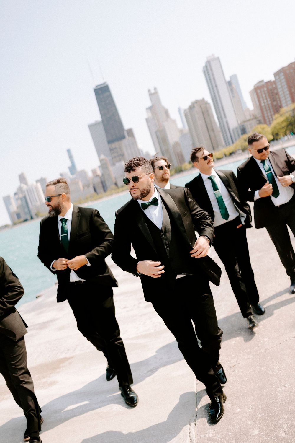 Groomsmen in black suits and sunglasses walk along Chicago's waterfront with the city skyline in the background.