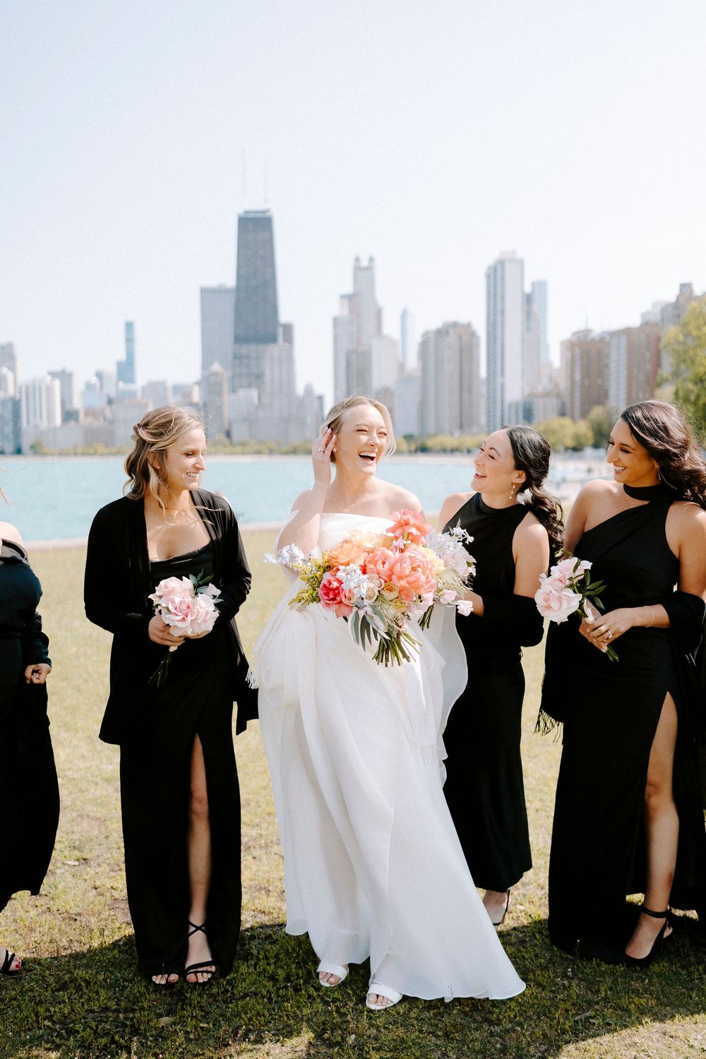 Wedding party in black dresses and white gown pose playfully against Chicago skyline backdrop.