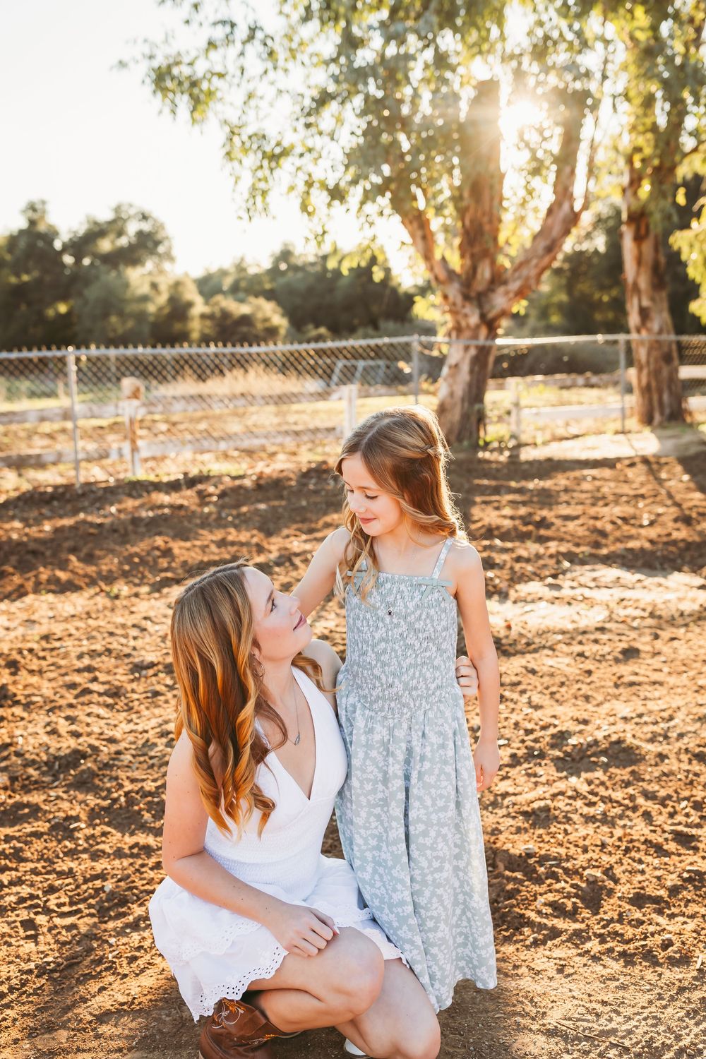 Mother and daughter share tender moments in golden sunlight on dusty ground beneath trees at sunset.