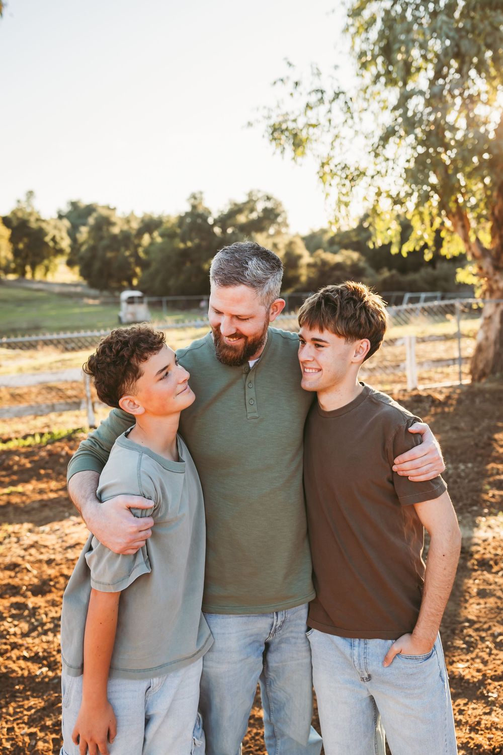 Three people embracing and laughing together outdoors during golden hour with trees in background.