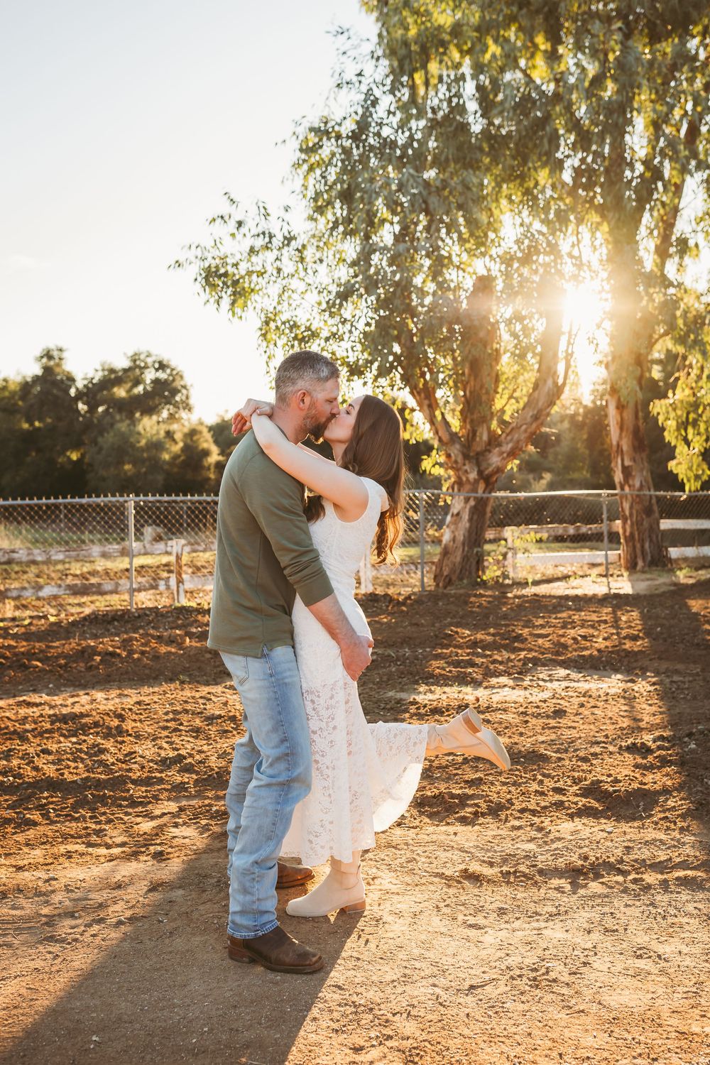 Romantic couple dancing and embracing in warm sunset light under tall trees in a rustic outdoor setting.