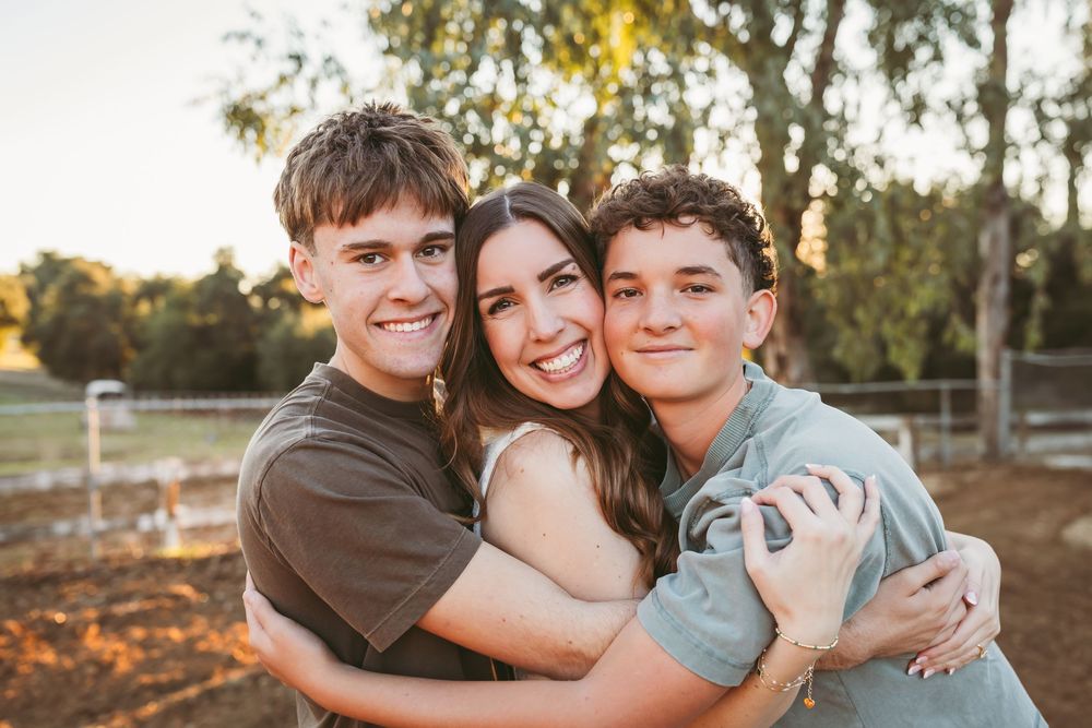 Three people embrace and smile warmly together outdoors during golden hour with trees in the background.