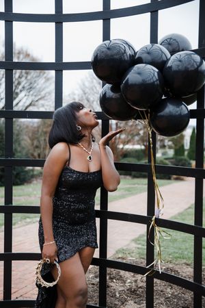 A woman in a black sequin dress poses with black balloons against a metal fence for a stylish photo shoot.