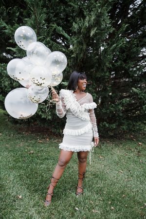 Someone in a white dress holds clear balloons filled with gold confetti while posing on grass near trees.