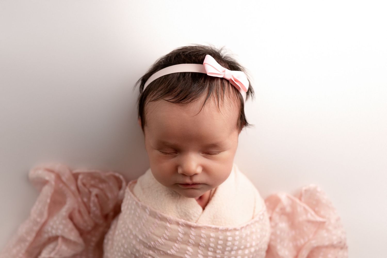 Sleeping newborn baby wrapped in soft pink swaddle with matching headband during professional photo session.