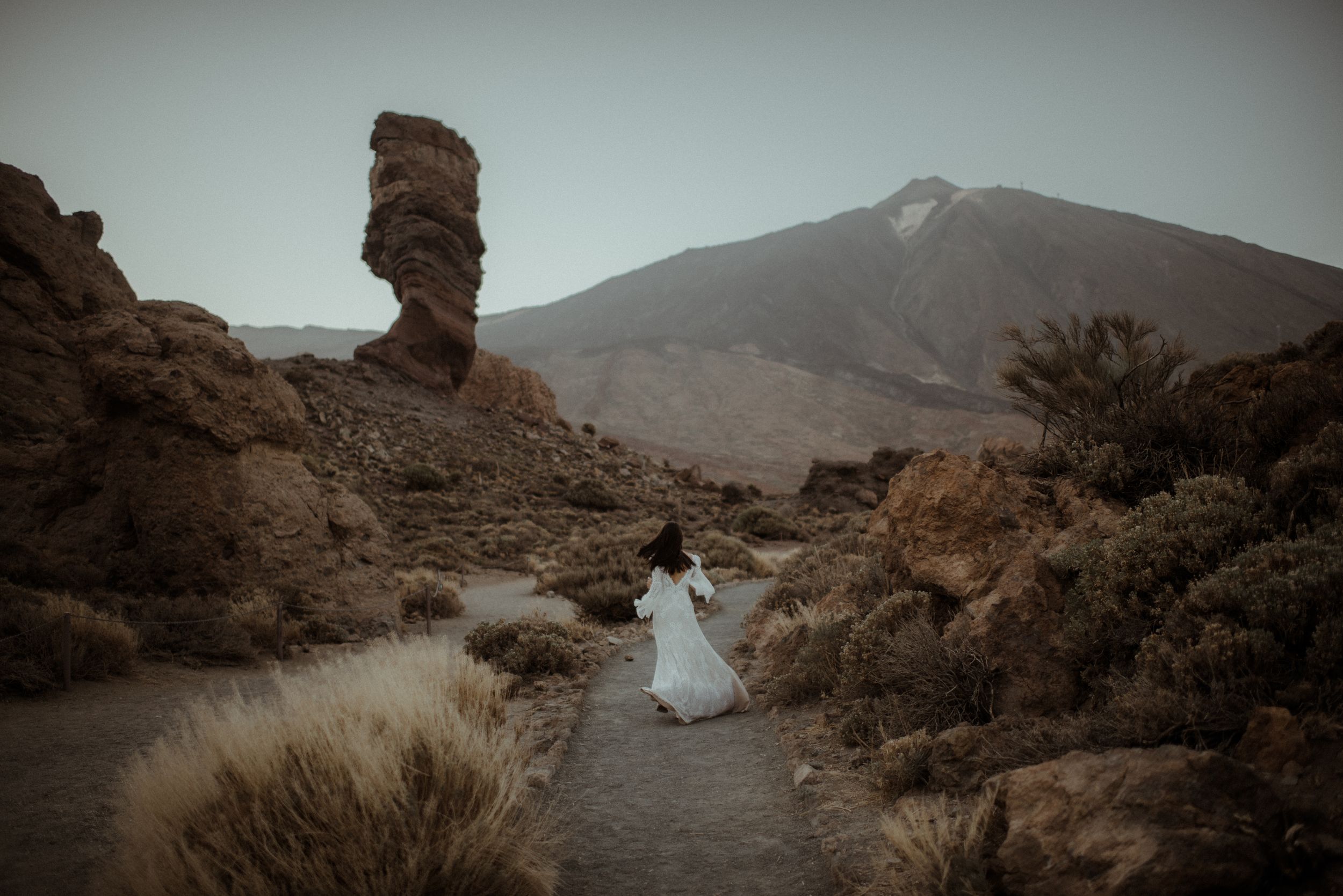 White lace dress flowing on a rocky mountain path with distinctive rock formation and mountain peak in background.