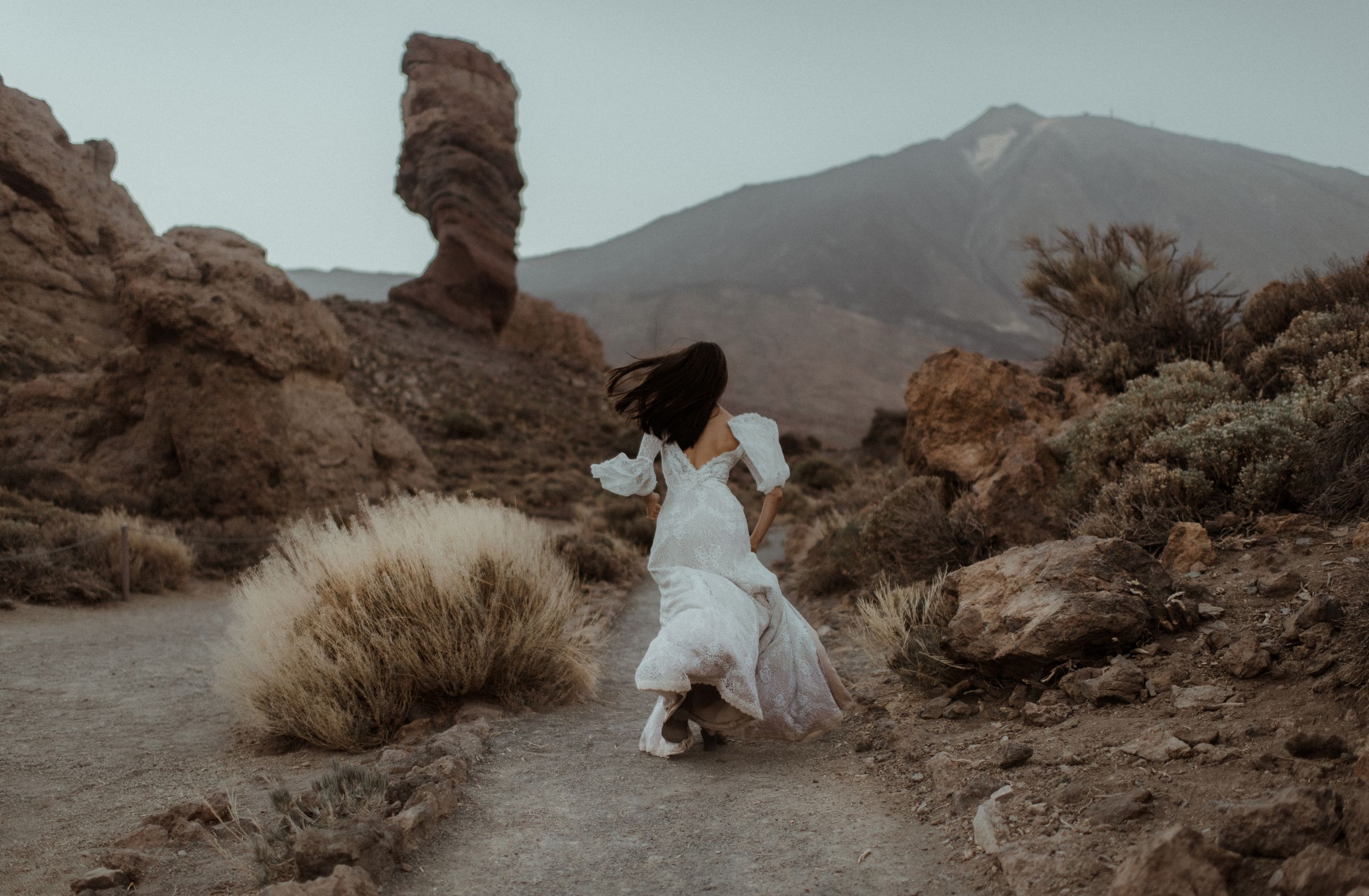 White wedding dress flowing dramatically on path near distinctive rock formation and mountain vista.