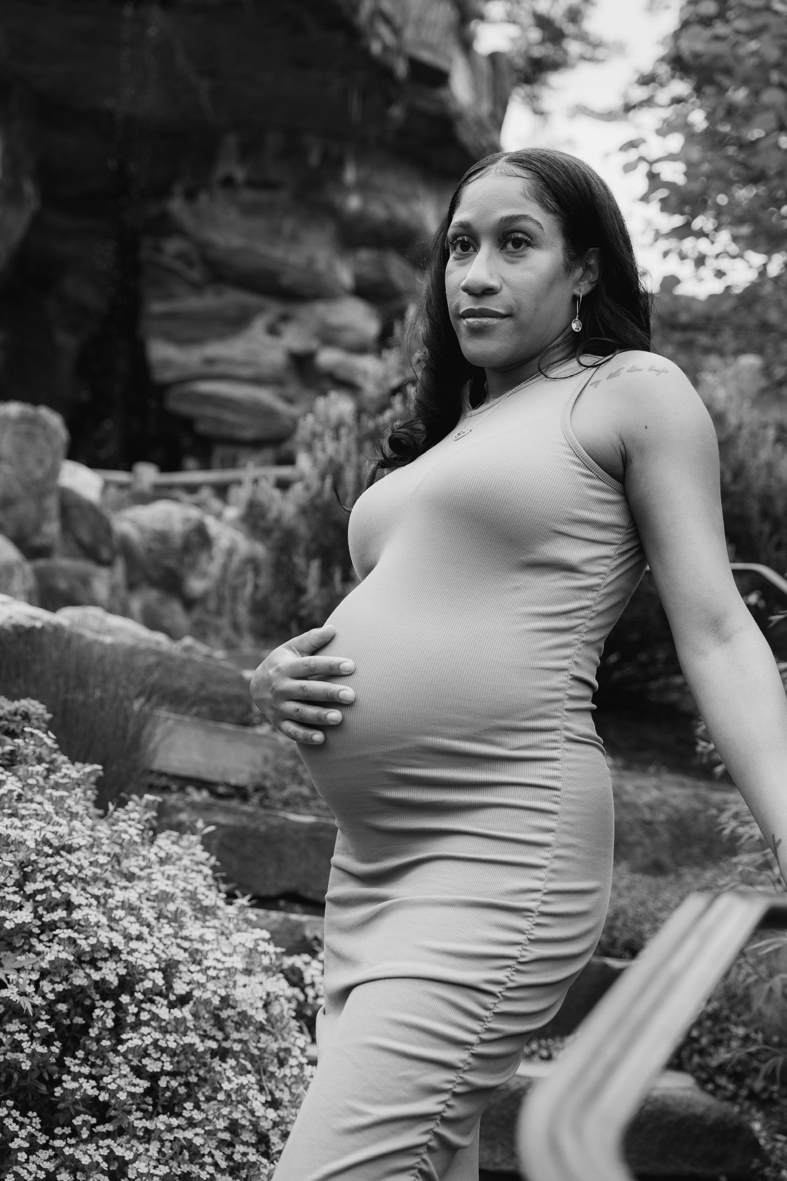 An expectant mother poses outdoors in a fitted dress against a rocky backdrop in black and white.