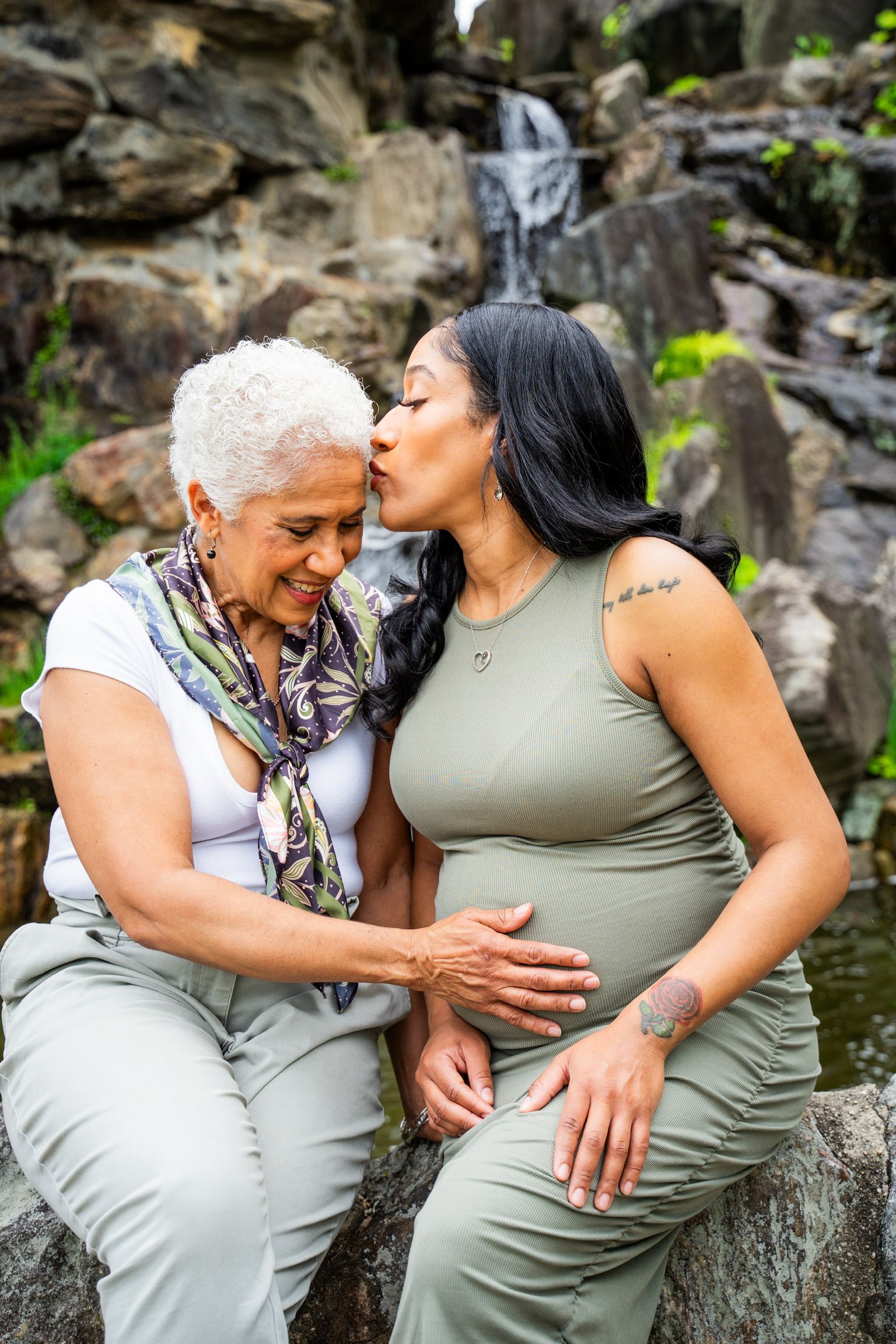Pregnant woman and older relative share touching moment by waterfall in natural setting.