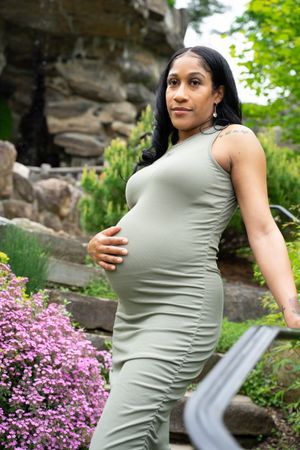Expectant mother in olive dress poses confidently against rocky garden backdrop with purple flowers.