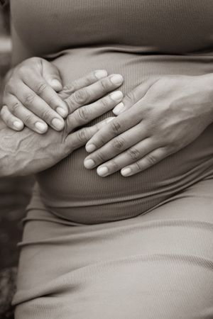 Pregnant woman in grey dress touching her baby bump while standing in a lush garden setting.