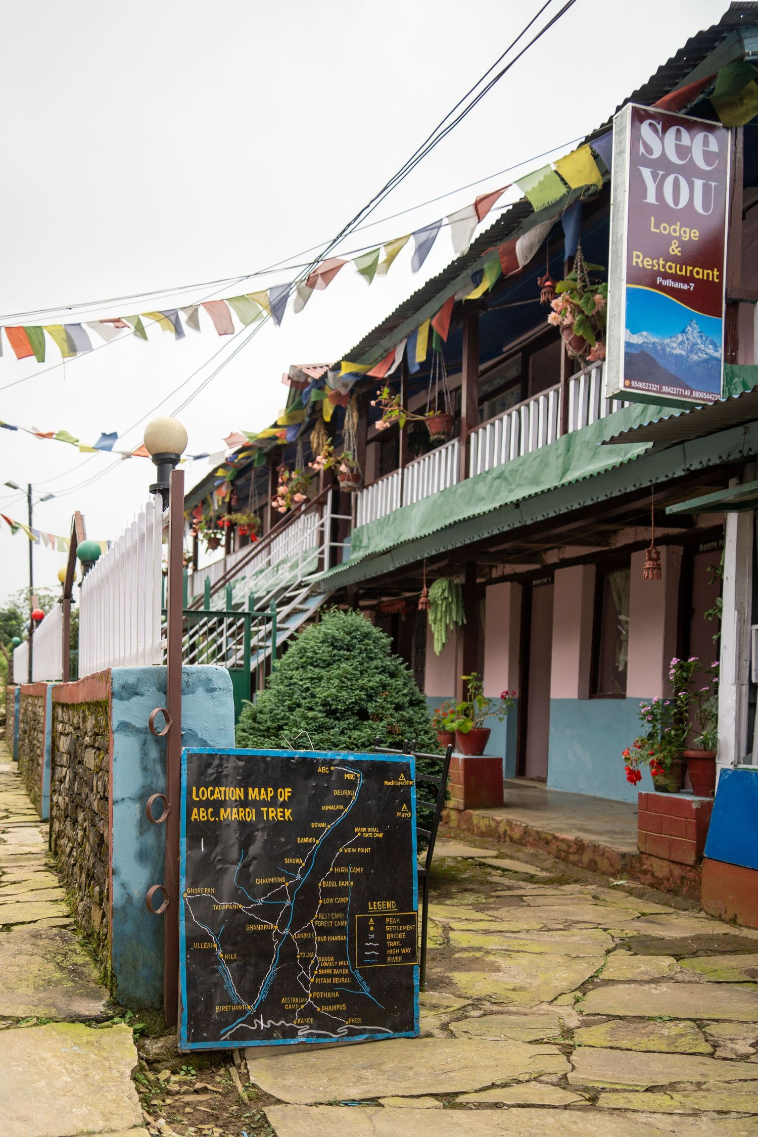 Mountain lodge buildings with colorful prayer flags and signage along a stone path.