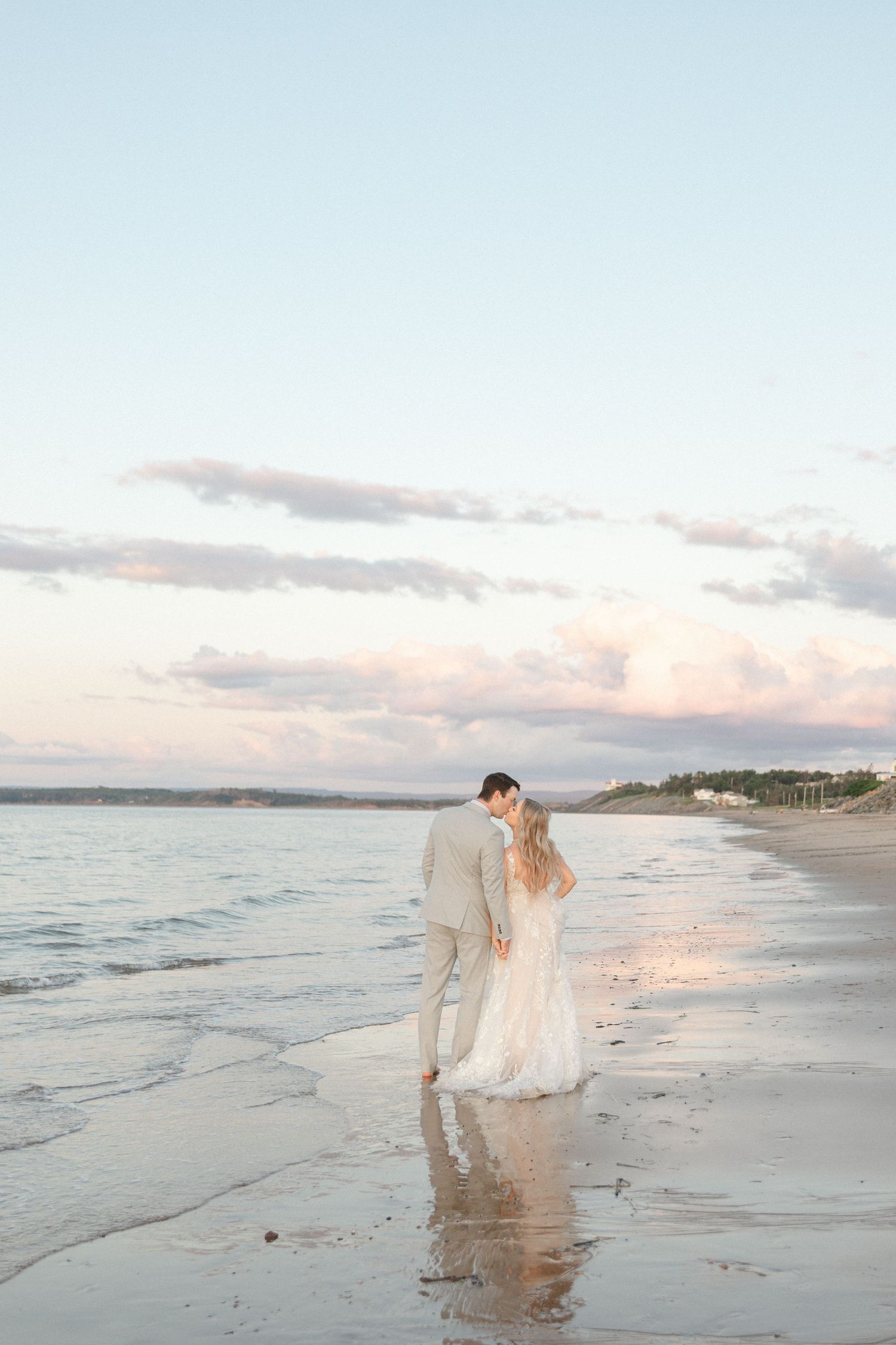 A couple walks along a serene beach at sunset with soft waves lapping at their feet while wearing elegant attire.