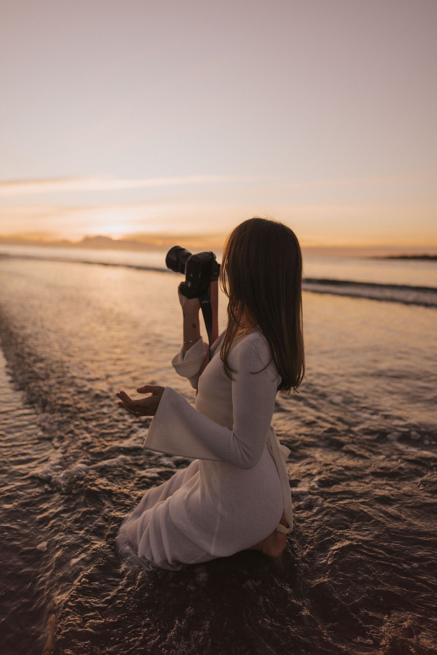 Woman in white dress kneeling on wet beach photographing ocean at golden sunset.