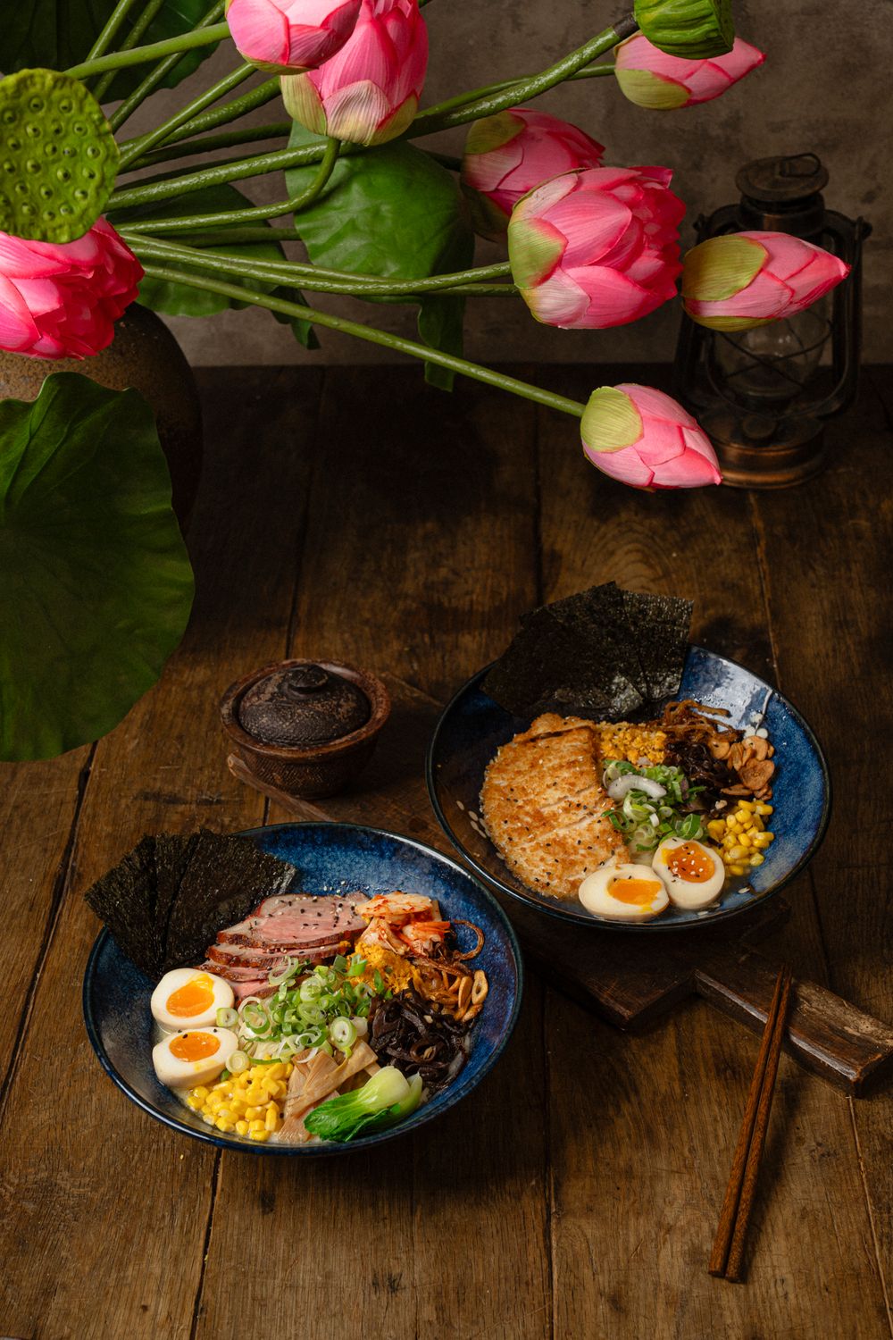 Two bowls of ramen with soft boiled eggs, meat and vegetables alongside pink tulips on wooden table.