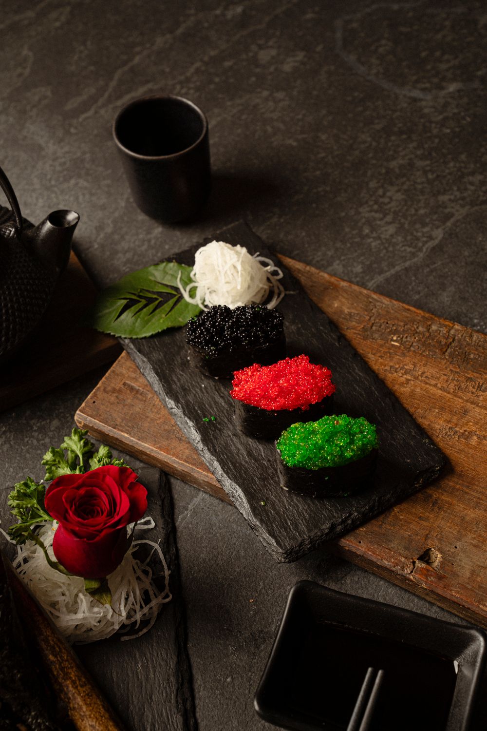 Japanese condiments and tobiko caviar displayed on slate with traditional teapot and red rose garnish.