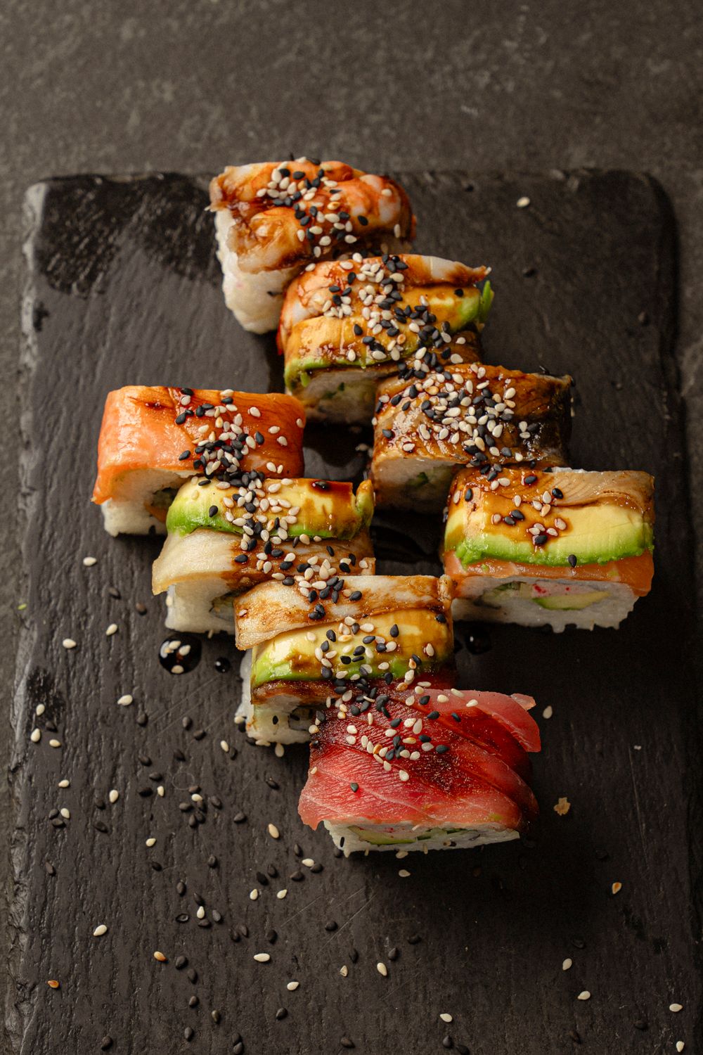 Artfully plated sushi rolls with salmon and tuna topped with sesame seeds on black slate.