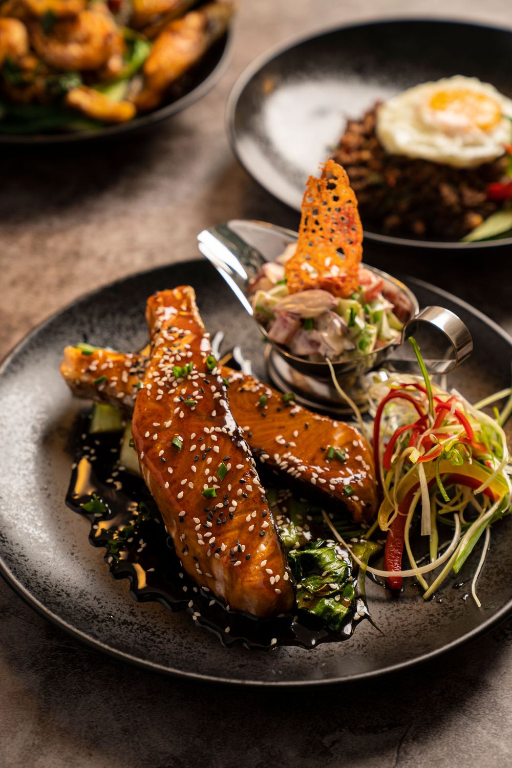 Glazed chicken wings topped with sesame seeds and served with fresh vegetables on a black plate.