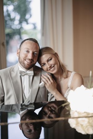 Smiling bride and groom pose together at a reflective table, groom in gray suit with bow tie, bride in white lace dress.