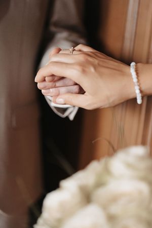 Bride and groom holding hands, showing wedding ring and pearl bracelet, with a white rose bouquet in the foreground.