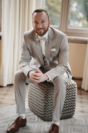Smiling groom in light gray suit with bow tie sits on woven ottoman near window in elegant wedding preparation setting.