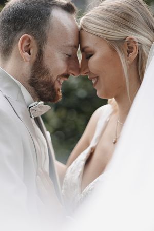 Smiling bride and groom touching foreheads tenderly on their wedding day, groom in gray suit, bride in white lace gown.
