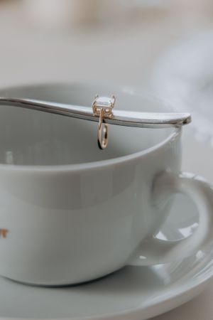 A delicate rose gold engagement ring sits perched on the rim of a white porcelain teacup and saucer.
