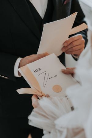 Hands holding wedding vows with "Her Vows" written on paper with wax seal and elegant ribbons.