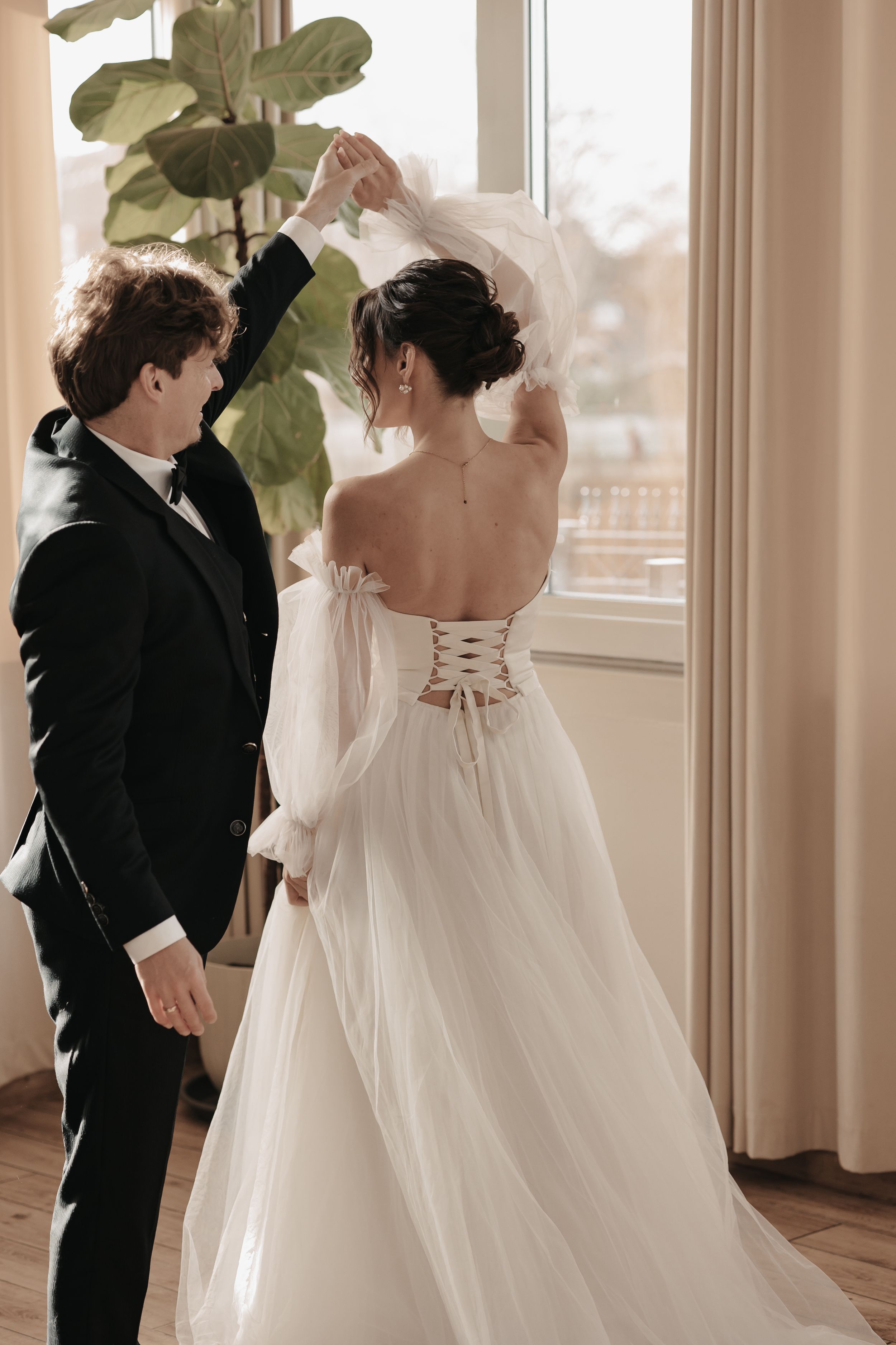 A bride in a flowing white corset gown with tulle skirt stands by a window with groom in classic black tuxedo.