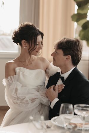 A tender romantic moment between newlyweds at their wedding reception, seated at an elegant table with wine glasses.