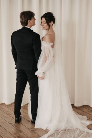 A romantic wedding photo of a couple in formal attire standing against a light backdrop with wooden flooring.