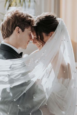 Romantic wedding portrait with gentle forehead touch between newlyweds under flowing white veil.